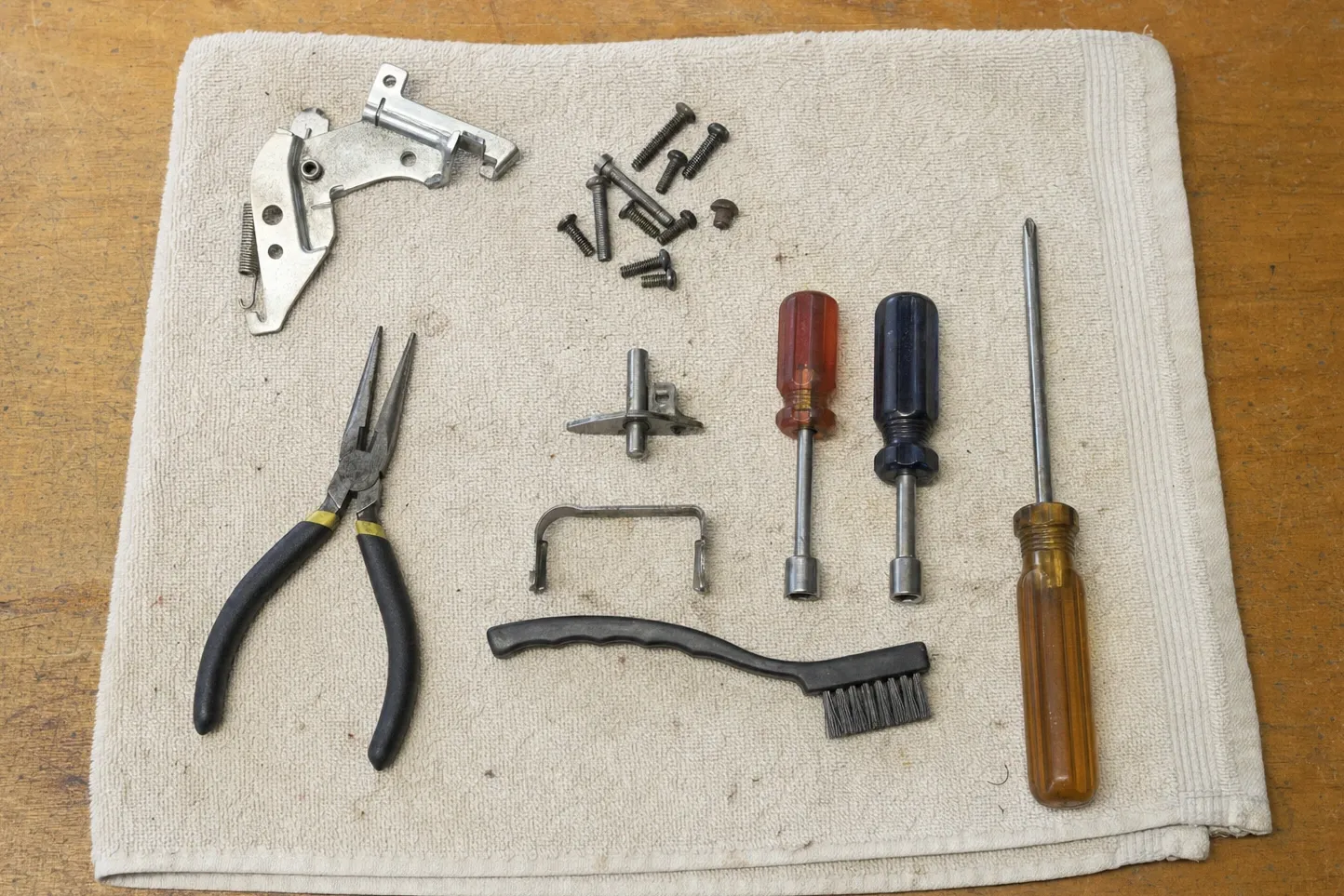 Oven door hardware and hand tools laid out on a cloth: hinge parts, a wire form, loose screws, drivers and a wire brush