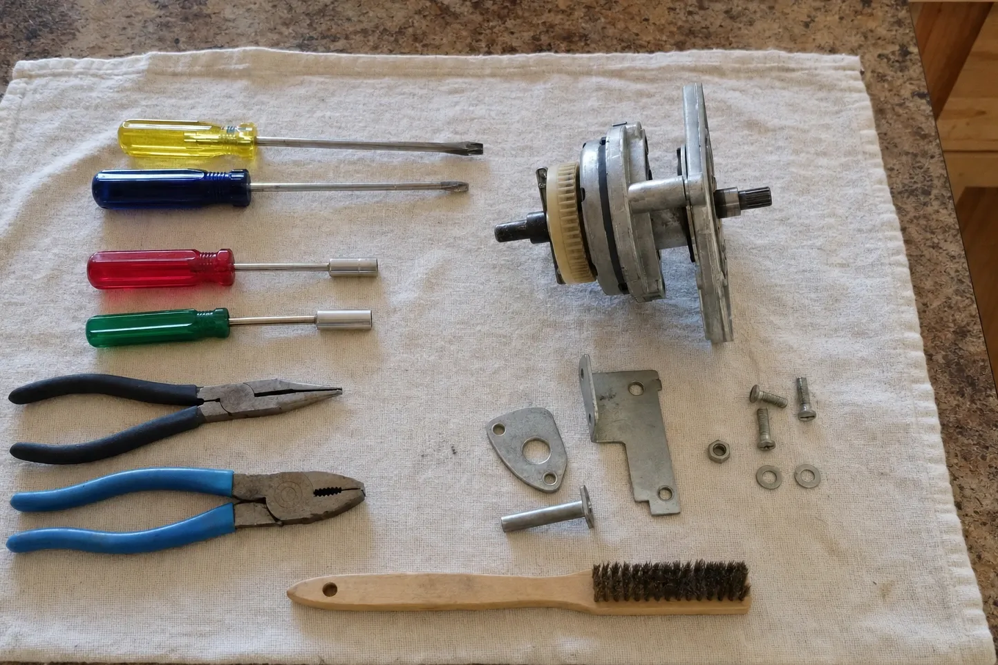 A removed motor with its shaft, plastic end-bell and mounting plate set on a cloth beside four colored drivers, two pairs of pliers and a wire brush