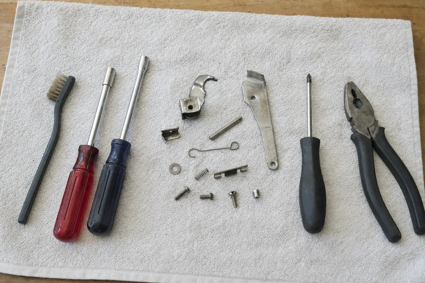 A curved cam lever, a flat strike plate, a hairpin clip, a light spring and a pivot pin on a gray towel with a wire brush, drivers and pliers